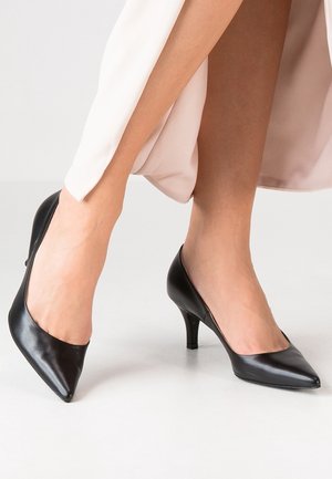 Woman wearing black pointed-toe mid-heel shoes paired with light-colored pants, standing on a smooth, light floor.