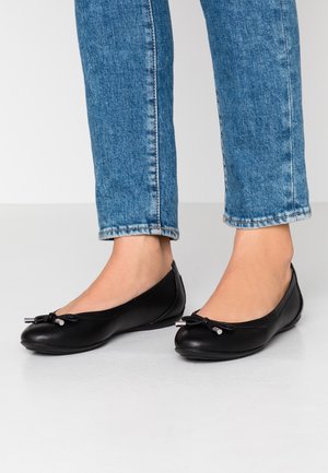 Füße in schwarzen Leder-Balletschläppchen mit kleinen Schleifen, kombiniert mit blauen Jeans, die über den Knöcheln enden, auf einem hellen Boden.