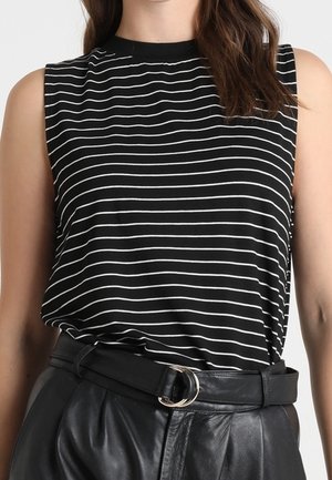 Vrouw draagt een mouwloze zwarte top met witte horizontale strepen, ingestopt in een zwarte leren broek met hoge taille en een dubbele ringriem.