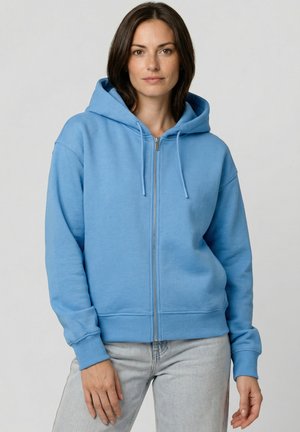 Frau trägt eine hellblaue Kapuzenjacke mit Reißverschluss, Kapuze und Kordeln, kombiniert mit hellgrauen Jeans, vor einem einfarbigen Hintergrund.