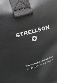 Čierna, textúrovaná taška s výrazným logom "Strellson" v bielej farbe. Obsahuje hashtagy a geografické súradnice vytlačené v bielej farbe.