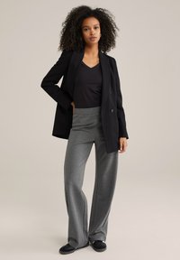 Femeie cu păr creț, îmbrăcată cu un sacou negru, un top negru cu decolteu în V, pantaloni gri largi și pantofi negri, stând cu o mână în buzunar.