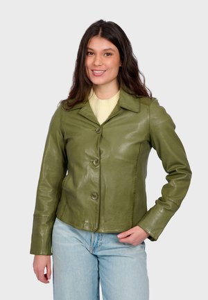 Žena se usmívá, má na sobě zapnutou olivově zelenou koženou bundu přes světlý svetr a světle modré džíny, s jednou rukou v kapse.