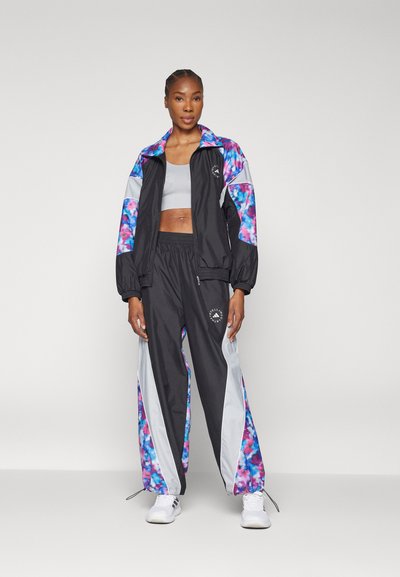 Mustat ja moniväriset juoksutakit, joissa on kukkakoristeet, yhdistettynä mustiin housuihin, joissa on harmaat ja kukkakuvioiset sivupaneelit. Valkoiset lenkkarit.