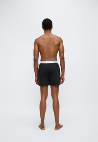 Μαύρα ανδρικά εσώρουχα boxer με λευκή ελαστική μέση με το λογότυπο Calvin Klein, εμφανιζόμενα από πίσω. Απλό σχέδιο, βαμβακερό υλικό, χωρίς πρόσθετα μοτίβα.