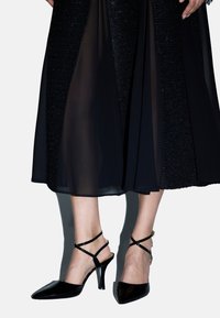 Scarpe col tacco a punta in pelle verniciata nera con design a cinturini, caratterizzate da un tacco sottile e cinturini alla caviglia, presentate su un tessuto scuro.