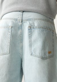 Vaaleansiniset farkut, joissa on kulunut viimeistely, kaksi takataskua ja hillitty keltainen ompelu taskulogossa.