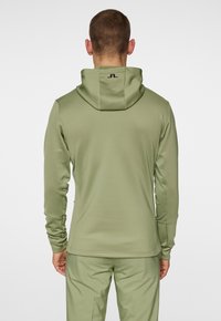 Vihreä urheilu hoodie, jossa on muotoiltu malli, sileä pinta ja huppu. Takana ja hihansuissa on minimaalista brändäystä.