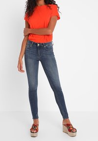 Camiseta roja de manga corta combinada con jeans ajustados azules, con un diseño clásico de cinco bolsillos. Sandalias de plataforma tipo cuña con acentos de patrón floral.