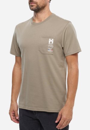 Homme portant un T-shirt à manches courtes brun clair avec une poche poitrine affichant le logo "Millet Guides Chamonix", assorti à un pantalon foncé.