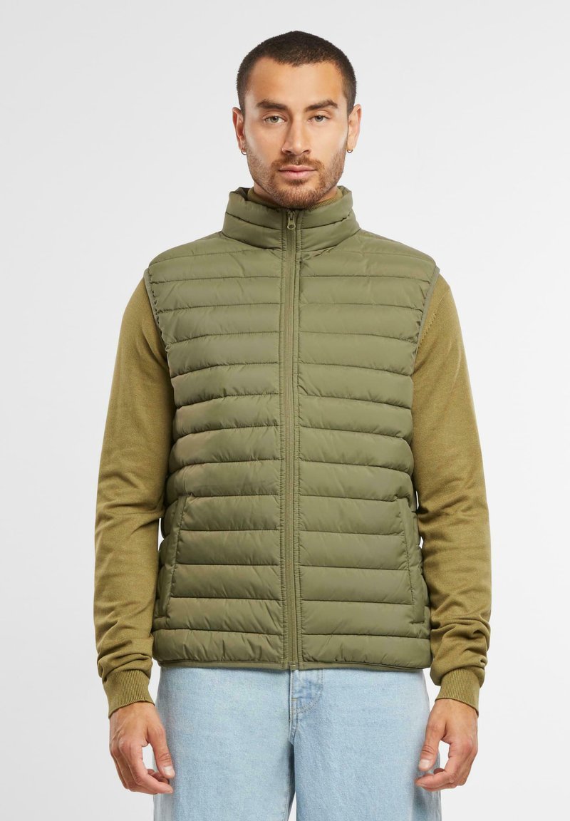 Olivgrüne Wattierte Weste mit hohem Kragen, horizontalen Nähten, Reißverschluss vorne, getragen über einem beigen Langarmshirt und hellblauen Jeans.