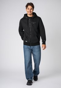Schwarzer Reißverschluss-Hoodie mit Kordelzügen, ausgestattet mit Fronttaschen und Logodetails. Kombiniert mit lockeren blauen Jeans und schwarzen Sneakers.