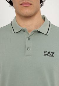 Light green polo shirt made of textured fabric, featuring a two-button placket, black collar accents, and a small EA7 logo on the chest.