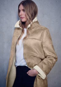 Chaqueta de ante sintético color marrón con cuello y puños de borreguito, cierre de cremallera y bolsillos laterales; llevada sobre una camiseta blanca y jeans oscuros.