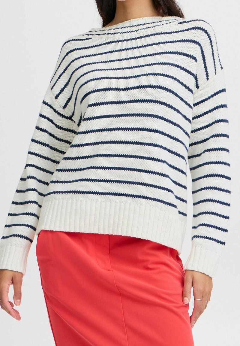 Femme portant un pull en tricot blanc à rayures horizontales bleu marine et un pantalon taille haute rouge vif, debout devant un fond blanc.