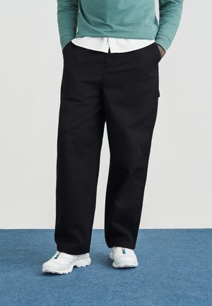 Persona con pantalones negros de pierna ancha, zapatillas blancas con detalles azules, una camisa blanca y un top verde de manga larga, de pie sobre una alfombra azul.