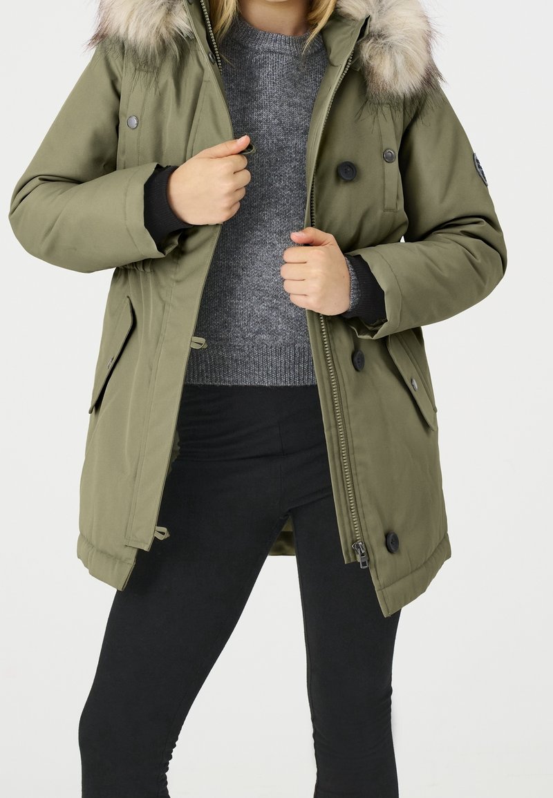 Personne portant un manteau d’hiver vert olive avec une capuche bordée de fourrure par-dessus un pull gris et un pantalon noir, tenant le manteau ouvert au niveau de la fermeture éclair.