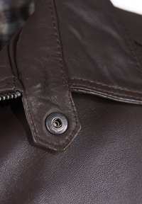 Nærbillede af en mørkebrun læderjakke med metaltrykknap og detaljeret syning nær lynlåsområdet.