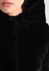 Černý kabát z umělé kožešiny s načechranou strukturou, kapucí a knoflíkovým zapínáním u krku. Látka vypadá měkce a silně.