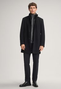 Cappotto di lana nera con colletto alto, due tasche frontali e chiusure a bottoni, indossato sopra una giacca a vento grigia e pantaloni neri slim.