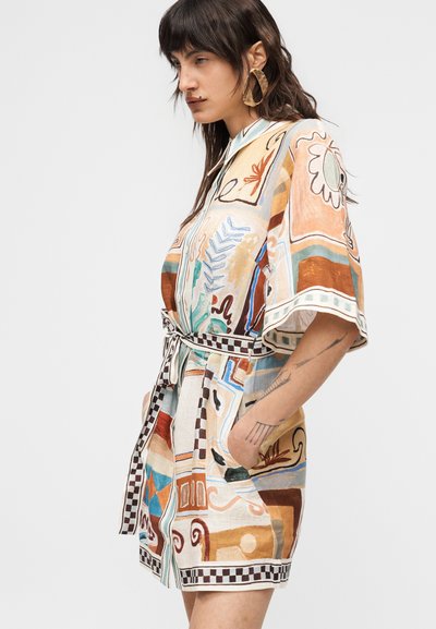 Γυναίκα με σκούρα μαλλιά μήκους ώμου, ντυμένη με φορεματικό με σχέδια, ζώνη στη μέση, φαρδιά μανίκια και μεγάλα χρυσά σκουλαρίκια, που ποζάρει πλαγίως με τα χέρια στις τσέπες.