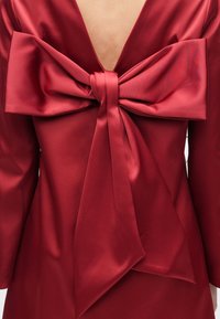 Bakfra av en person som har på seg en rød satengkjole med en stor, elegant sløyfe knyttet nederst på ryggen og en dyp V-formet ryggutringning.