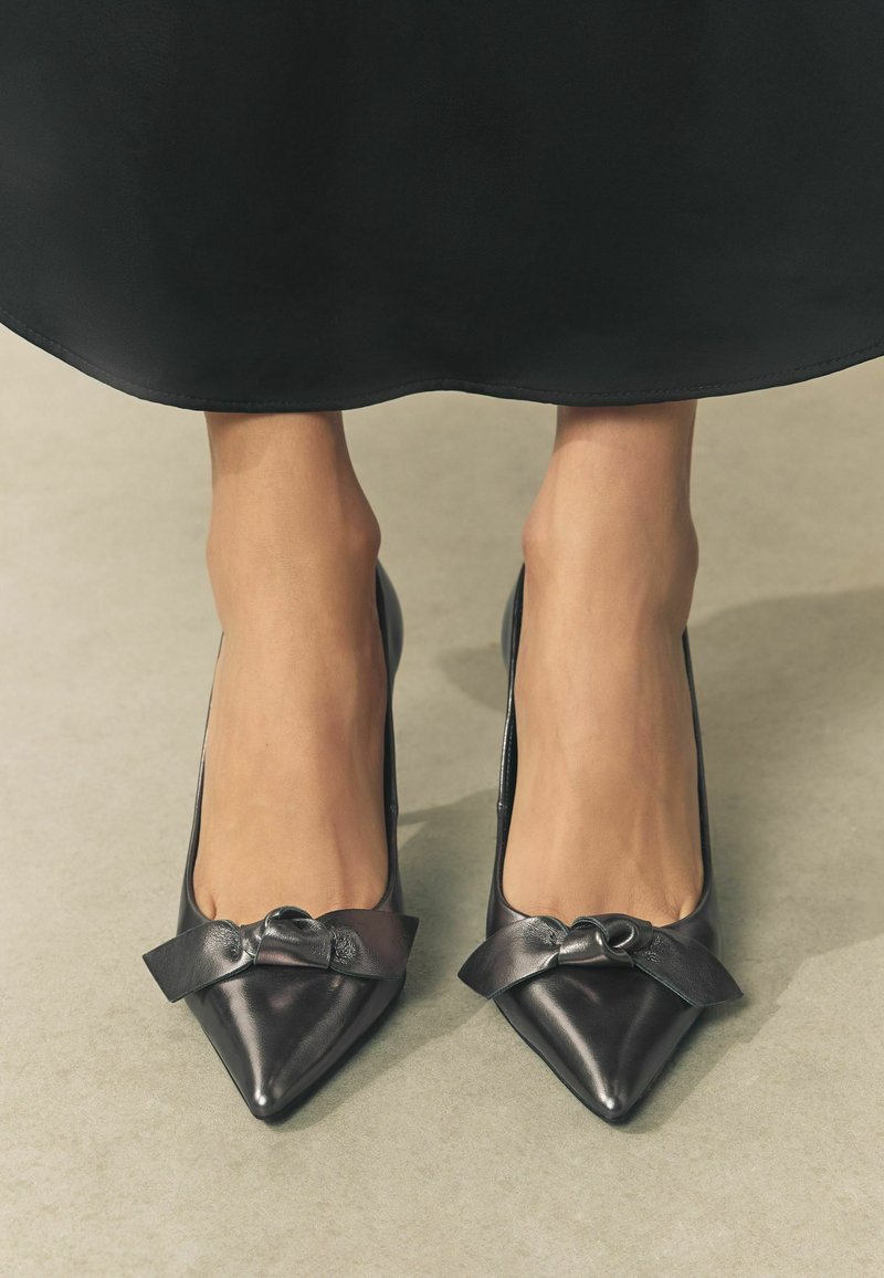 Schwarze Leder-Pumps mit spitzem Zehenbereich und einem dekorativen Schleifenakzent an der Vorderseite. Glatte Textur mit glanzvollem Finish.