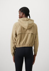 Kvinne med flettehår iført en beige kortermet hettegenser og svarte leggings, stående mot en enkel hvit bakgrunn, vendt bort.