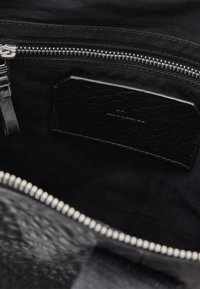 Interior de una bolsa negra con una etiqueta de cuero texturizado y cierre de cremallera plateado, con un forro de tela negra suave y herrajes en contraste.