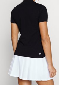 Person som bär en svart kortärmad polotröja med texten "ULTRA DRY" och en vit veckad kjol, sedd bakifrån.