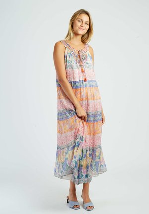 Kobieta stojąca, ubrana w bezrękawnikową, wielokolorową kwiecistą sukienkę maxi i jasnoniebieskie buty na otwartym obcasie na jednolitym białym tle.