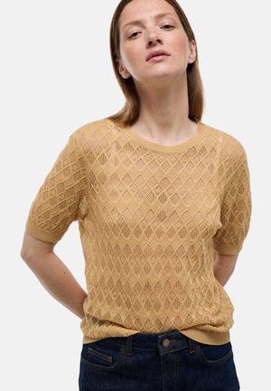 Femme portant un haut en maille beige à manches courtes avec motif losange et un jean bleu foncé, debout les mains dans le dos, fond neutre.