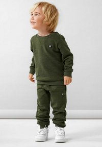 Felpa in pile verde oliva e pantaloni jogger con dettagli in logo nero, abbinati a sneakers bianche. Texture morbida e vestibilità rilassata.