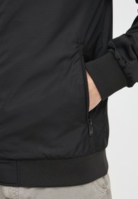 Person der bærer en sort jakke med ribbede manchetter og kant, hånd inde i den lynlåsede sidelomme, beige bukser synlige ved taljen.