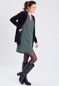 Cardigan en maille noire sur une robe à motifs verts, accompagnée de bottes noires jusqu'aux genoux. La robe a une coupe décontractée et des manches courtes.