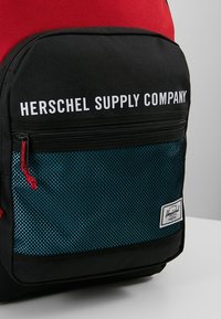 Sac à dos noir et rouge avec une poche avant en maille couleur sarcelle, texte du logo en blanc, fermeture éclair noire et une étiquette de marque blanche en bas à droite.