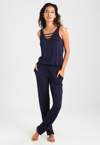 Der marinefarbene Jumpsuit verfügt über einen V-Ausschnitt mit drei horizontalen Trägern, einem elastischen Taillenbund, ärmellosem Design und seitlichen Taschen für zusätzlichen Komfort.