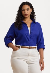 Camisa de linho azul royal com botões à frente e mangas arregaçadas, combinada com calças brancas de cintura alta e um cinto castanho.