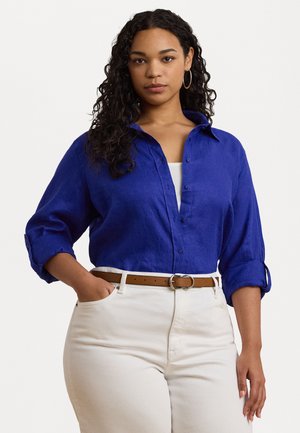 Koninklijkblauwe linnen blouse met een knoopsluiting aan de voorkant en opgerolde mouwen, gecombineerd met hooggetailleerde witte broek en een bruine riem.