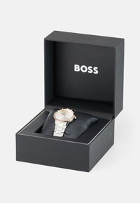 Silberne Metall-Uhr mit rosegoldenem Ziffernring, weißem Ziffernblatt und verlinktem silbernen Armband, präsentiert in einer schwarzen Box mit Logo.
