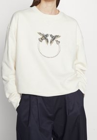 Crämfärgad sweatshirt med ett dekorativt tryck av två fåglar och ett cirkulärt mönster. Tillverkad av mjukt tyg med en avslappnad passform.