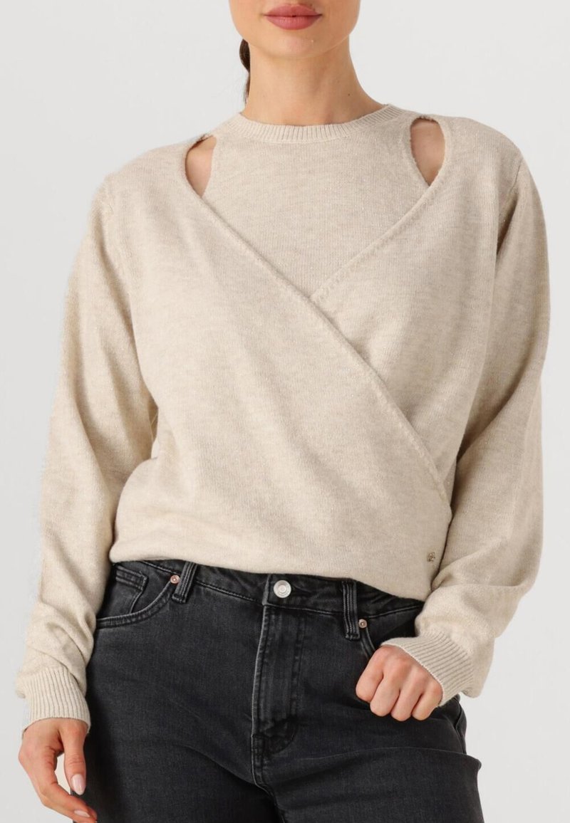 Beige trui met een crossover ontwerp, voorzien van open schouderuitsparingen en geribde manchetten. Gecombineerd met donkere spijkerbroeken.