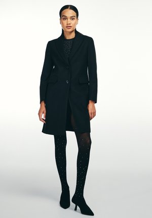 Cappotto nero su misura con un singolo bottone, colletto a risvolto e tasche laterali, abbinato a collant neri trasparenti decorati con piccole embellishments argento.