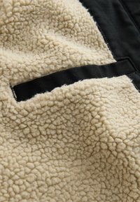 Tela de forro polar texturizada beige con un panel y detalles de bolsillo en negro contrastante. Superficie suave y esponjosa con un patrón ligeramente elevado.