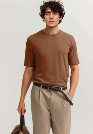 Mann mit lockigem Haar, der ein braunes Kurzarmshirt und beige Hose mit geflochtenem Gürtel trägt, und eine braune Tasche in einer Hand hält.