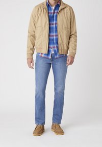 Kratší bomber bunda přes modrou kostkovanou košili, spárovaná se světle modrými džíny a hnědými slip-on botami. Hladká tkanina, casual design.