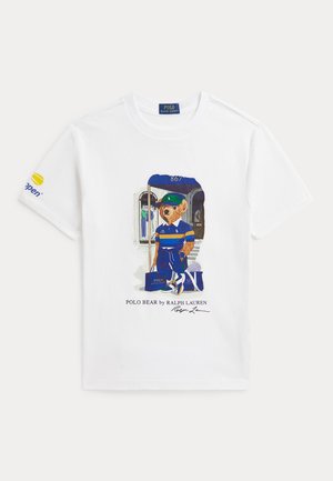 Hvid t-shirt med grafik af en bjørn iført blå sportstøj, der holder et flag og tennisketsjere, med teksten Polo Bear by Ralph Lauren nedenunder.