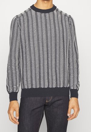 Homme portant un pull en tricot rayé verticalement noir et blanc avec des poignets, un ourlet et un col côtelés, associé à un jean foncé.