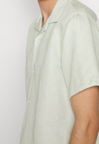 Camicia verde chiaro a maniche corte con bottoni e motivo circolare sottile. Il materiale sembra leggero e leggermente texturizzato.