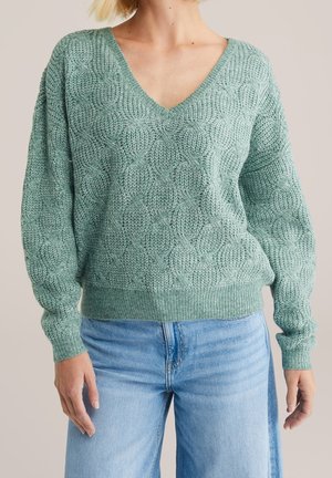 Femme portant un pull en maille texturée vert à col en V avec des manches longues et un jean bleu clair sur un fond uni.